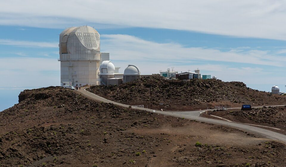 Haleakala_Observatory_2017_ Ett stort astronomiskt observatorium med flera vita teleskopkupoler ligger på ett kargt, mörkt lavafält på en bergstopp. En smal väg slingrar sig mellan byggnaderna under en blå himmel med tunna moln.