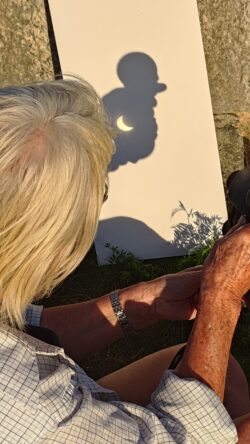 IMB and binocular-projected partial eclipse_20260812 En person med gråblont hår och rutmönstrad skjorta sitter i solen och håller i en kikare. På en vit skiva syns skuggan av kikaren med en tydlig halvmåneformad projektion av en partiell solförmörkelse.