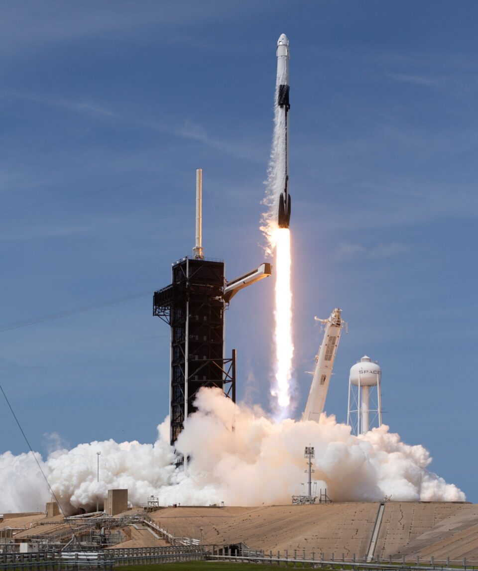 SpaceX_Demo-2_Launch from KSC_public domain En vit SpaceX Falcon 9-raket lyfter från en uppskjutningsramp och lämnar efter sig en ljus eldkvast och ett tjockt moln av vit rök mot en klarblå himmel.