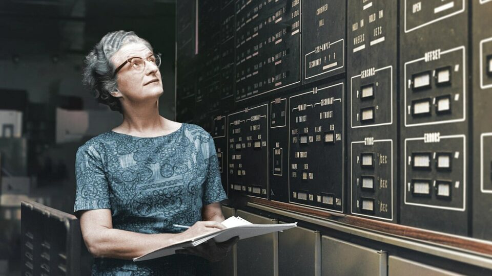 Nancy_Roman_Hubblecast_NASA_CC BY 2-0 En äldre kvinna står självsäkert framför en vägg fylld med datorskåp, i en teknologisk miljö.