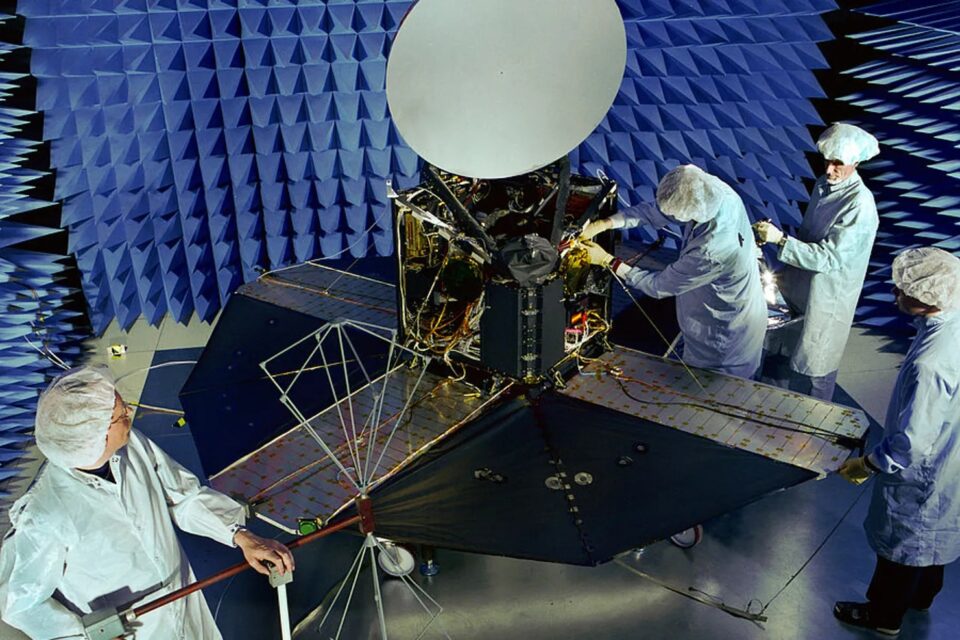 Odin_Rymdstyrelsen Tekniker i renrumsdräkter arbetar med en satellit utrustad med solpaneler och en stor antenn, inne i ett rum klätt med blå, ljudabsorberande skumkilar.