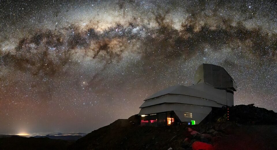 Rubin_Observatory_and_Its_Target_CC BY 4.0 En vit astronomisk observatoriebyggnad står på en mörk, klippig bergskam under en färgstark, stjärnfylld Vintergata på natthimlen.