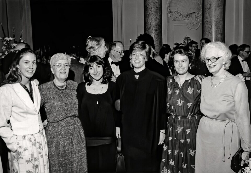 Several_women_scientists_who_changed_astronomy-CC BY 4.0 Svartvitt gruppporträtt av sex kvinnor som ler vid en formell mottagning, med elegant klädda gäster som minglar i ett praktfullt inrett rum.