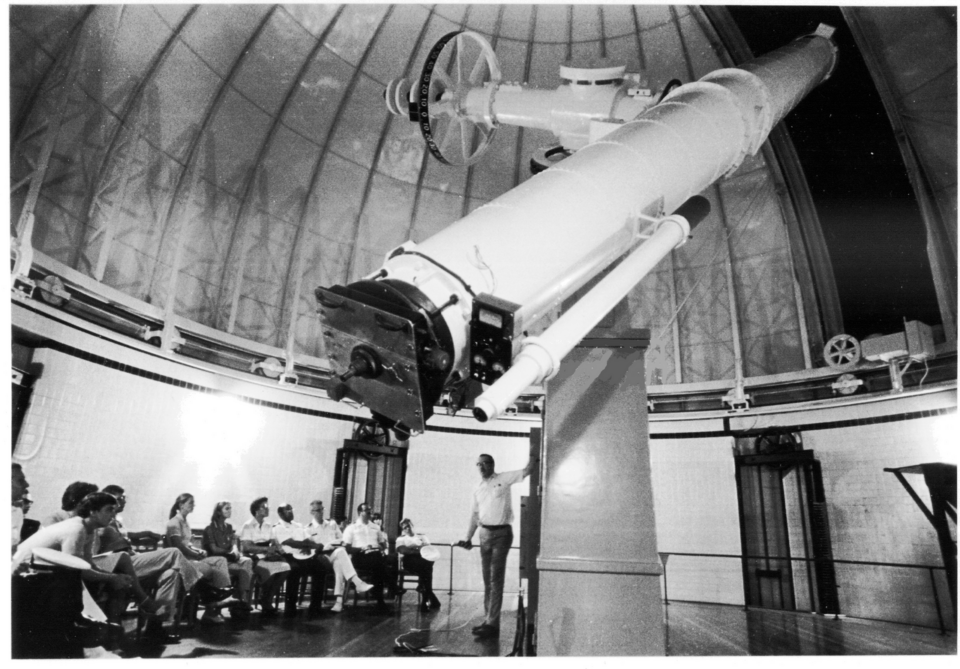 USNO 26-inch Clark telescope_public domain En man står bredvid ett stort teleskop inne i ett kupolformat observatorium och håller en presentation för en sittande grupp människor i ett svartvitt fotografi.