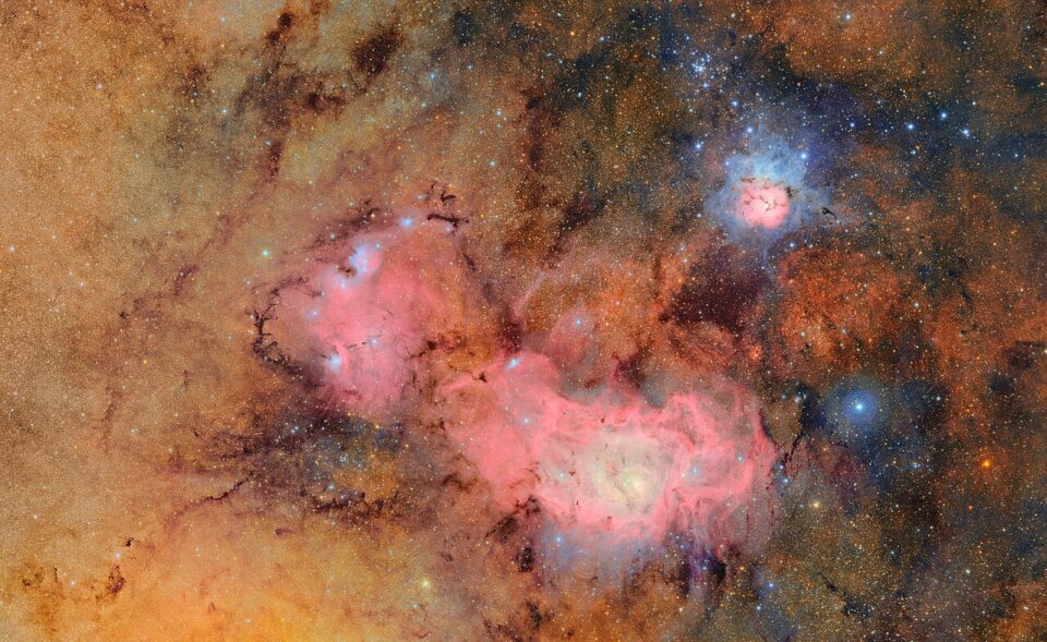 VRO 1st released picture_Trifid_and_Lagoon_nebulae_CC BY 4.0 En livfull vy över rymden med glödande rosa emissionsnebulosor mot ett tätt, gyllenbrunt stjärnfält med spridda, klarblå stjärnor.