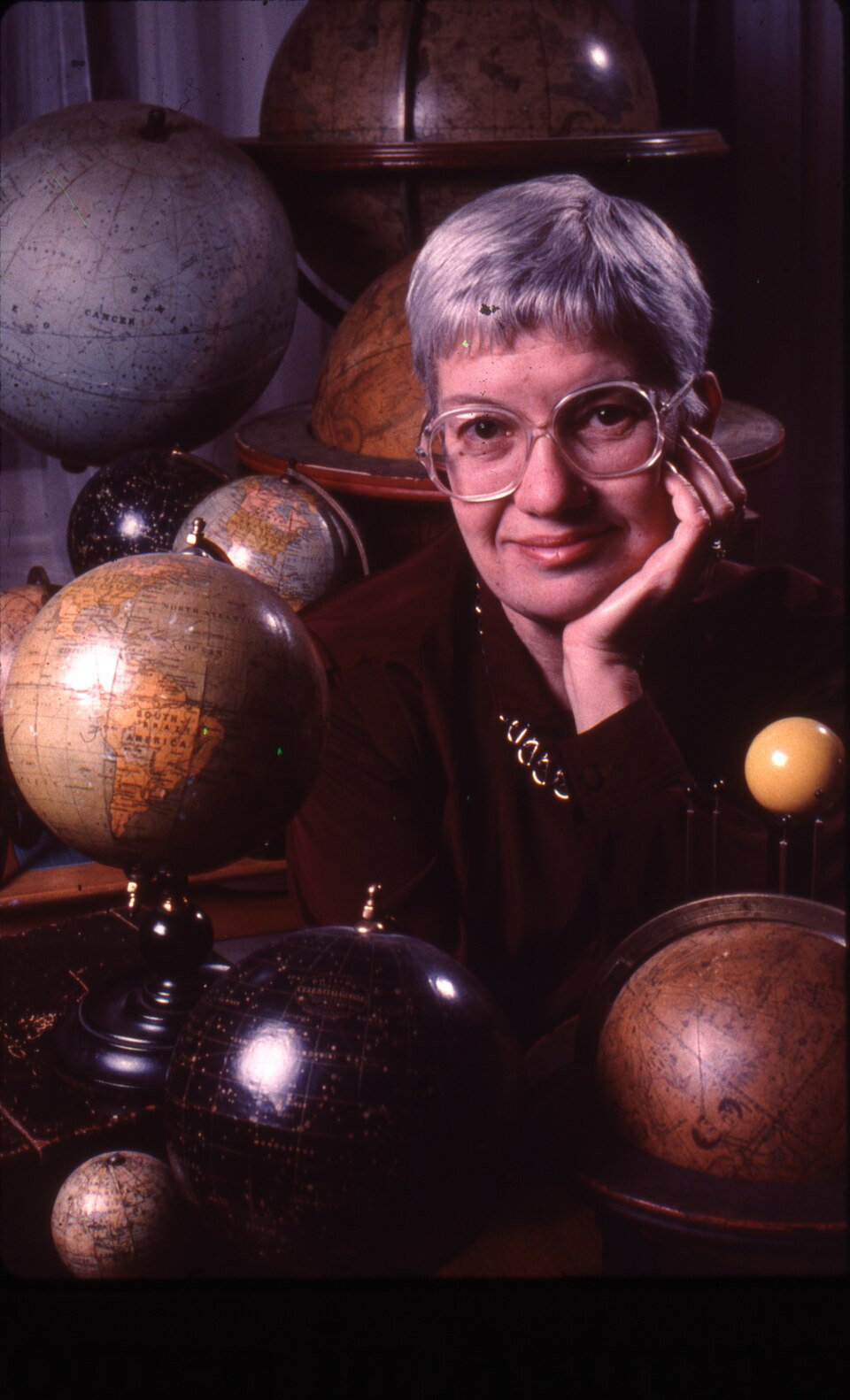 Vera_Rubin_with_antique_globes Porträtt av en leende kvinna med glasögon som vilar hakan i handen, omgiven av gamla jordglober i en varm, akademisk miljö.