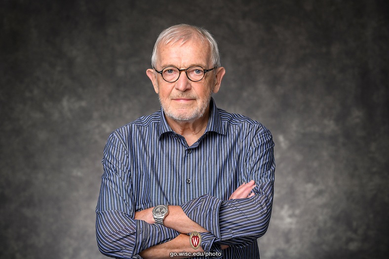 Francis Halzen_2026 Nobel in Physics_Althea Dotzour University of Wisconsin–Madison Bilden visar en äldre stilig man med glasögon och blå skorta med armarna i kors, poserar sittande framför en grå vägg.