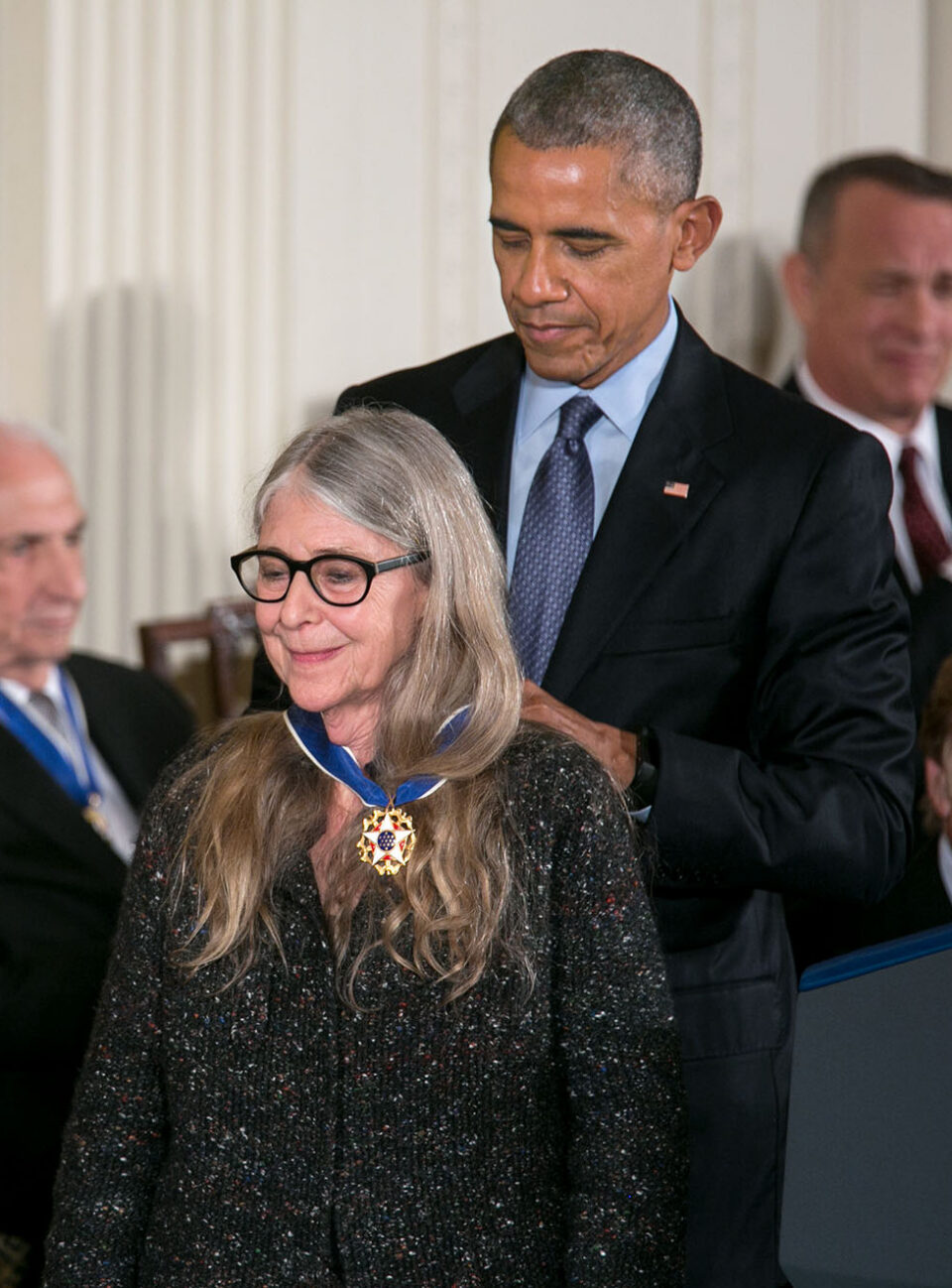 Hamilton_Medal_of_Freedom_from_Obama_public domain Barack Obama hänger presidentens frihetsmedalj runt halsen på Margaret Hamilton, som bär svartbågade glasögon och en mörk tröja.
