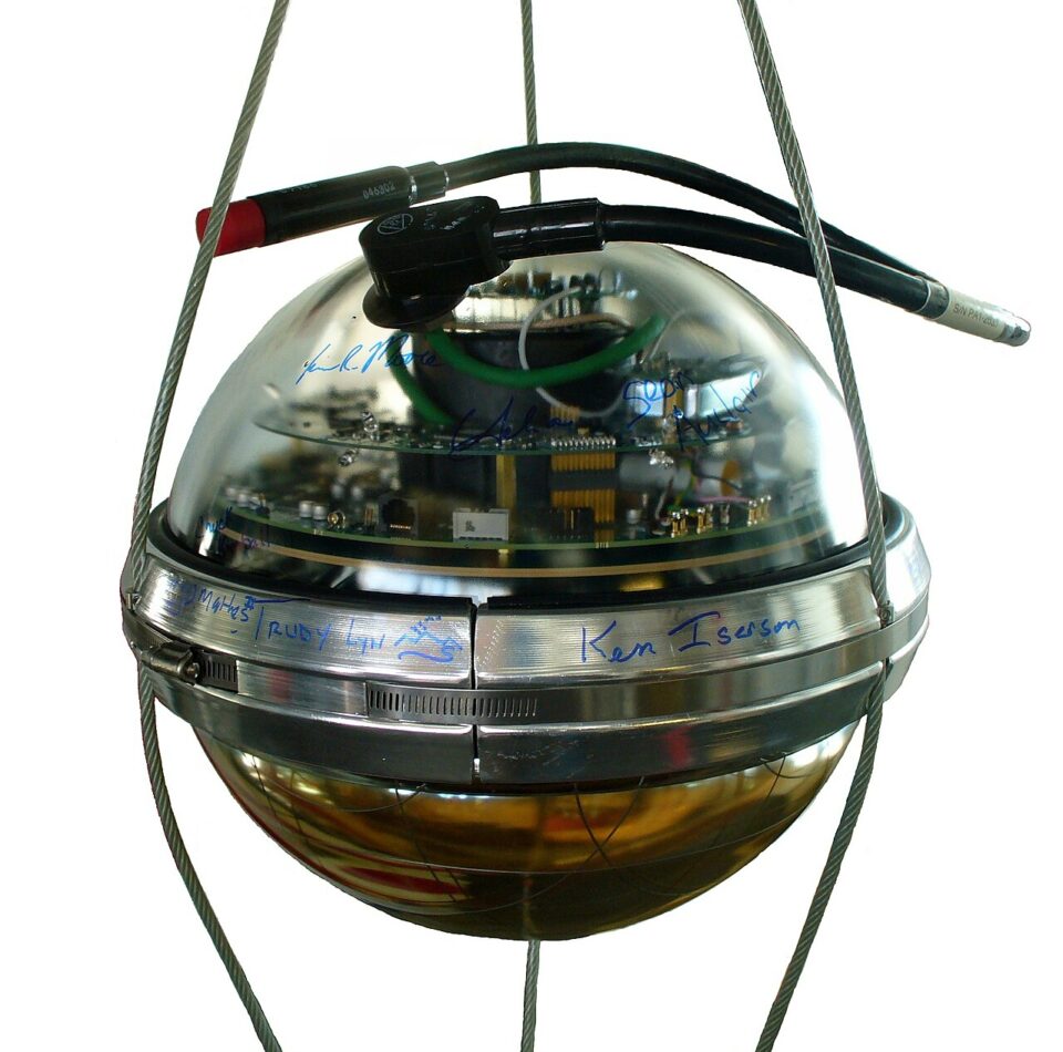 IceCube digital optical module-DOM-taklampa_Amble CC BY-SA 4.0_deployed in 2000 hole 85 Ett sfäriskt vetenskapligt instrument av glas och metall som hänger i vajrar, med interna kretskort, svarta kablar och handskrivna signaturer runt en metallkant på sfärens mitt.