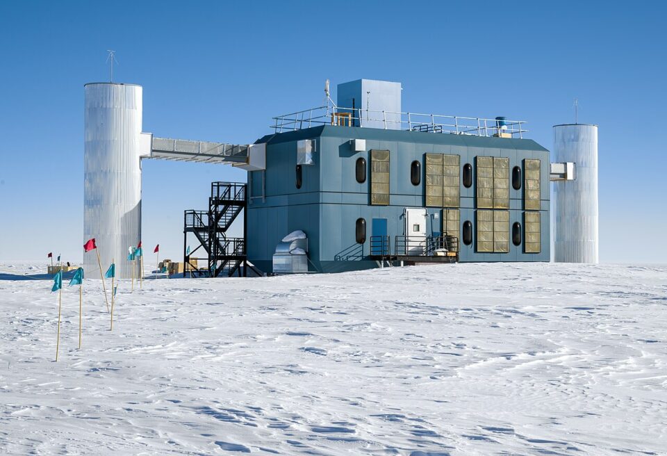 IceCube_Neutrino_Observatory_in_2023_Christoper Michel_CC BY-SA 4.0 En modern, blå forskningsstation ligger i ett vidsträckt, snöklätt antarktiskt landskap under en klarblå himmel, flankerad av två höga, cylindriska silvertorn.