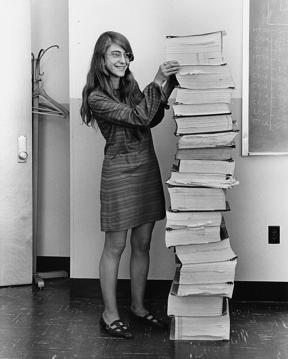 Margaret_Hamilton_-_restoration_public domain En leende kvinna med långt hår och glasögon står bredvid en mycket hög trave av tjocka, inbundna tekniska manualer i ett kontor med en vägg som är en griffeltavla.